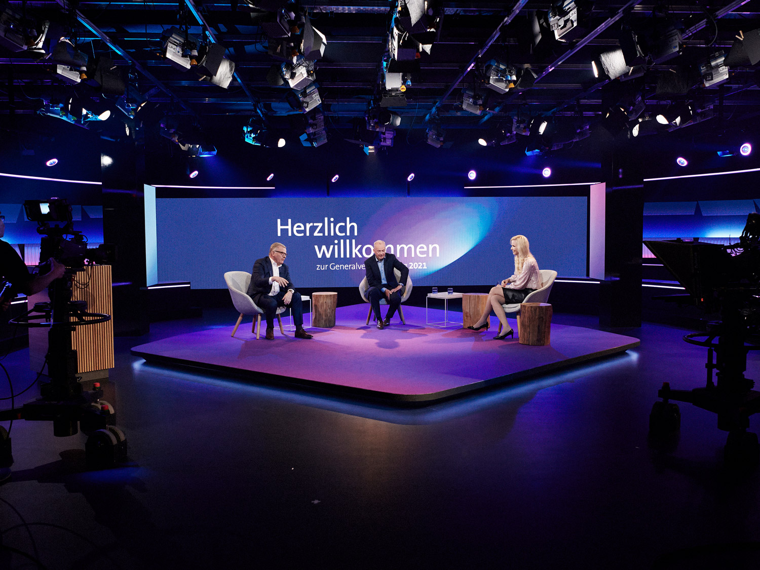 Drei Personen sitzen mittig im Bild auf einer Bühne. An der Decke sind Scheinwerfer montiert, im Hintergrund ist eine Leinwand um die Bühne sieht man Kameras.