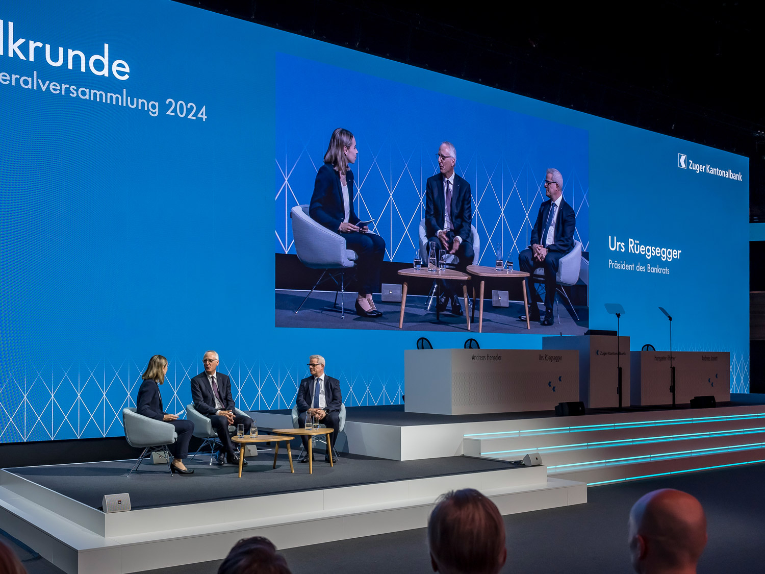 Bühnensituation an einer Generalversammlung mit drei Personen auf der linken Seiten des Bildes in einer Talk-Runde.