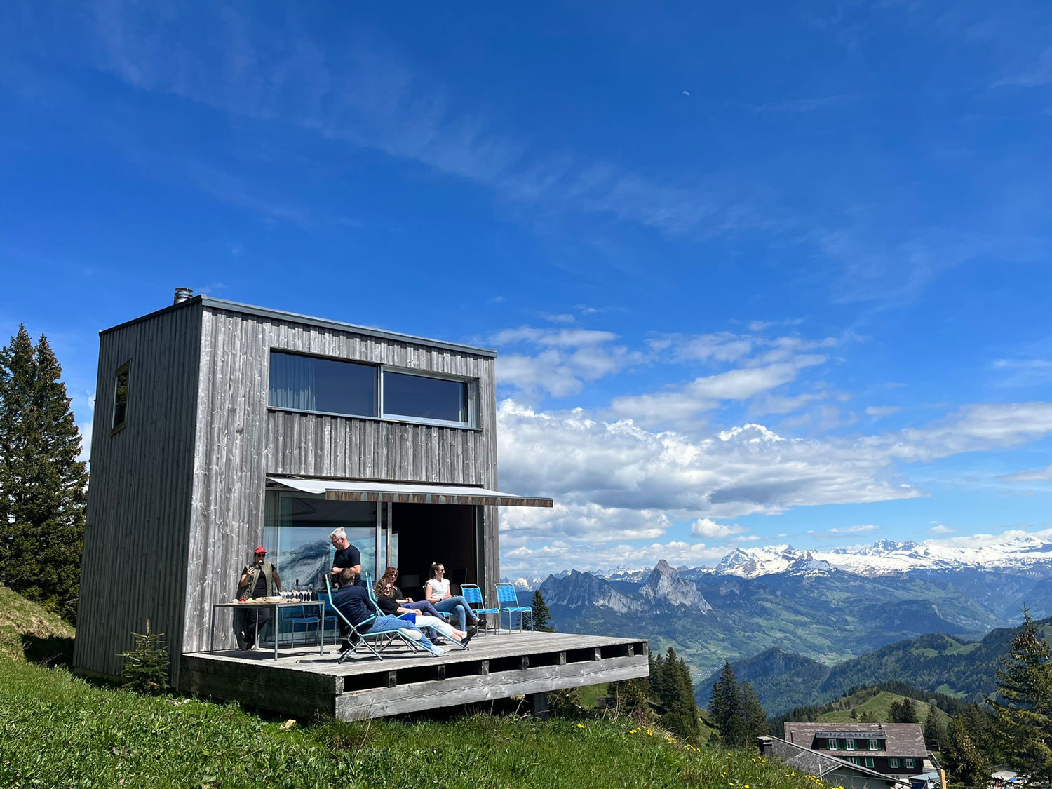 Haus mit Holzfassade und Berglandschaft im Hintergrund. Mehrere Personen sitzen auf der Terasse davor.